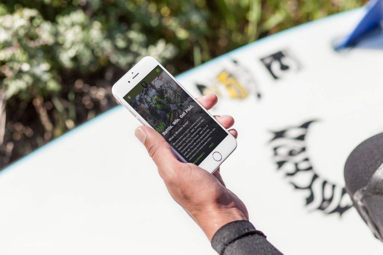 An athlete holding a phone showing the Monster Army app outdoors, beside a graphic-wrapped board.